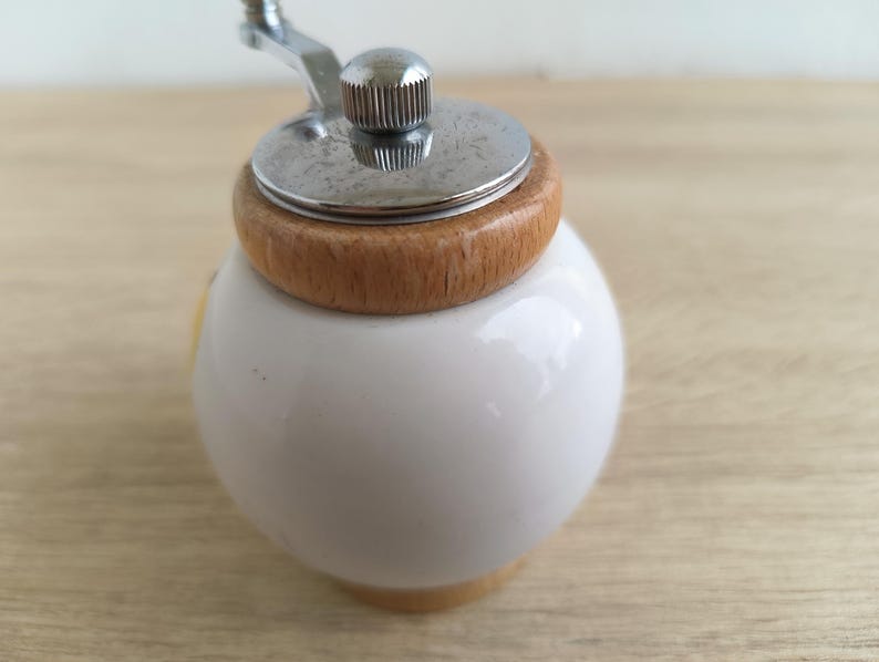 A small, round vintage-style pepper mill with a white porcelain body and a light wooden top. The mill features a subtle yellow geometric graphic on its side and has an exposed metal grinding mechanism at the top. The image is a close-up, showcasing the mill's design and internal structure, resting on a rustic farmhouse wooden table.