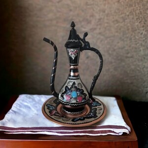 May include: A black and silver decorative teapot with floral designs and a matching tray. The teapot has a spout and a handle. The tray is round and has a raised edge.