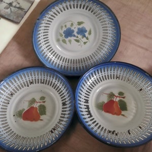 Puede incluir: Tres platos de metal vintage con interior blanco, bordes azules y diseño calado. Dos platos presentan una ilustración de pera roja, mientras que el otro tiene flores azules. Ideal para la decoración de la cocina.