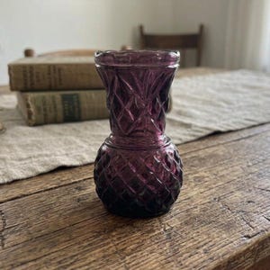 Puede incluir: Un pequeño jarrón de cristal amatista de estilo vintage con un patrón de diamantes. El jarrón tiene una base bulbosa y una parte superior acampanada. Se encuentra sobre una mesa de madera rústica, con libros y un camino de mesa de color neutro en el fondo.