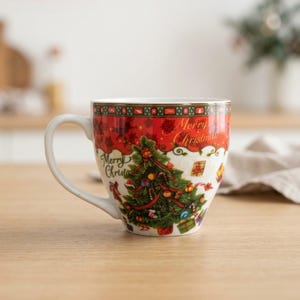 Könnte beinhalten: Ein weißer Keramikbecher mit einem weihnachtlichen Tannenbaum-Motiv, Geschenken und dem Schriftzug Merry Christmas in goldener Schrift. Der obere Rand ist mit einem rot-grünen Muster verziert. Der Becher hat einen geschwungenen Henkel.