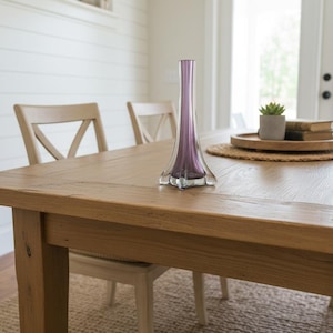 May include: A purple glass vase with a flared base stands on a wooden table. The vase has a long neck and a star-shaped base. The table is made of light-coloured wood and is surrounded by chairs. The table is approximately 150 cm long.