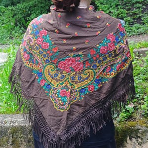 May include: A brown shawl with a floral design in red, pink, blue, and yellow. The shawl has a fringe trim and is draped over the shoulders. The shawl is a traditional accessory.