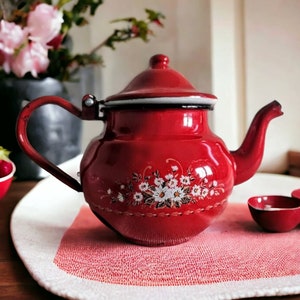 May include: A red enamel teapot with a white floral design on the side. The teapot is sitting on a red and white placemat.
