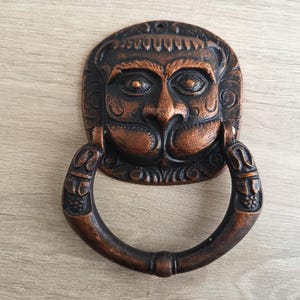 May include: Bronze-colored door knocker featuring a detailed face design with a ring handle. The knocker has an aged appearance, with intricate carvings around the face and handle. The face has a prominent nose and detailed eyes.