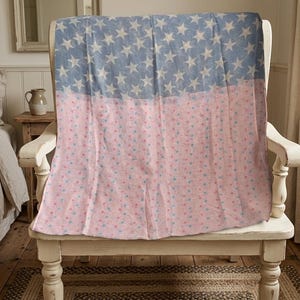 Pode incluir: Um cobertor azul claro e rosa com um padrão de estrelas e pontos drapeado sobre uma cadeira branca. A metade superior do cobertor apresenta estrelas brancas sobre um fundo azul claro, enquanto a parte inferior é rosa com pequenos pontos.