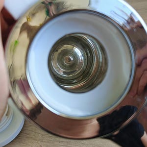 May include: Close-up of a shiny, chrome-finished object with a circular design. The object has a white interior ring and a central glass component. The reflective surface shows distorted reflections.