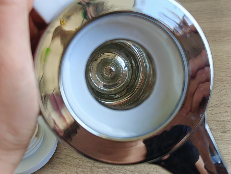 May include: Close-up of a shiny, chrome-finished object with a circular design. The object has a white interior ring and a central glass component. The reflective surface shows distorted reflections.