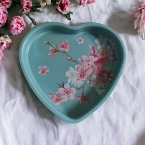 Puede incluir: Un molde para hornear de cerámica en forma de corazón con un glaseado azul claro y un diseño floral con flores de cerezo rosas y blancas.