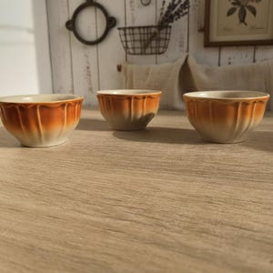 Könnte beinhalten: Drei kleine Keramikschalen mit einem gewellten Design. Die Schalen haben eine Farbverlaufsoptik, die von einem hellen Beige am Boden zu einem warmen Orange am Rand übergeht. Sie sind auf einer hellen Holzoberfläche angeordnet.