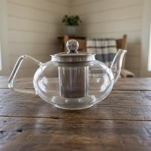 Op de afbeelding: Een heldere glazen theepot met een roestvrijstalen infuser en deksel. De theepot heeft een gebogen handvat en tuit. Hij staat op een rustieke houten tafel. De achtergrond omvat ramen en een glimp van een stoel.