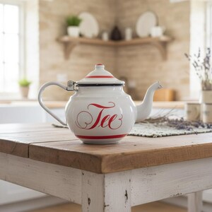 May include: White enamel teapot with a red stripe and the word "Tee" in red script. The teapot has a curved handle and a spout. It sits on a wooden table in a kitchen setting.