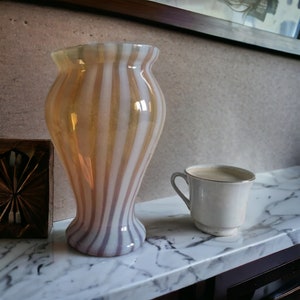 Op de afbeelding: Een witte en gouden gestreepte glazen vaas met een brede opening en een smalle basis. De vaas staat op een wit marmeren oppervlak naast een witte koffiekop.