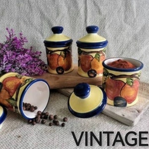 Puede incluir: Cuatro tarros de especias de cerámica vintage con diseños pintados en azul y amarillo de frutas y hojas. Los tarros están sobre una superficie de madera con la palabra "VINTAGE" impresa debajo.