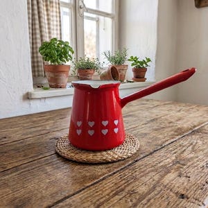 May include: A red enamel coffee pot with a long handle and white heart-shaped designs. The pot sits on a woven coaster atop a rustic wooden table. Potted herbs are visible in the background near a window.