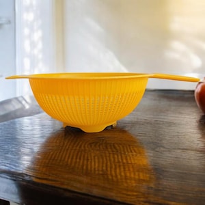 Puede incluir: Un colador de plástico amarillo con asa. El colador está sobre una mesa de madera.