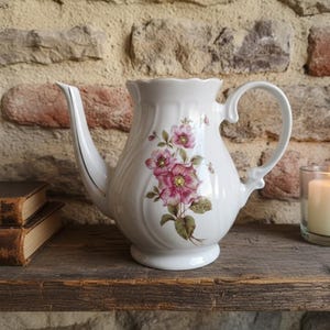 May include: A white ceramic teapot with a floral design in pink and green. The teapot has a curved spout and handle. A lit candle in a glass holder and two antique books are also visible.