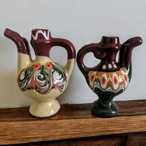 Puede incluir: Dos pequeñas teteras de cerámica con intrincados diseños pintados. Una es marrón con detalles blancos y amarillos, la otra es crema con detalles verdes y marrones.