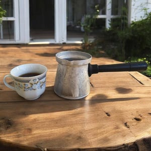 Puede incluir: Una cafetera de plata con mango negro y una taza blanca con un diseño floral sobre una mesa de madera.