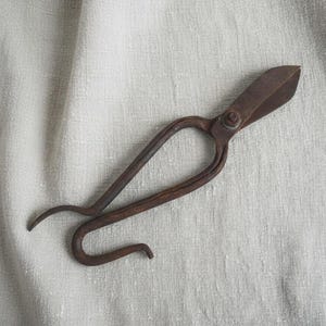 May include: Antique brown metal shears with curved handles and a scissor-like blade. The shears are resting on a light-colored, textured fabric. The blades are slightly open, suggesting they are ready for use.