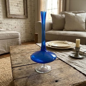 Könnte beinhalten: Eine leuchtend blaue Glasvase mit bauchigem Boden und schlankem Hals. Die Vase steht auf einem klaren, kreisförmigen Sockel. Sie steht auf einem rustikalen Holztisch, zusammen mit einer Kerze und Tellern.
