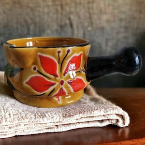 以下が含まれることがあります： 黒い取っ手のある小さな茶色の陶器のボウル。ボウルは赤い花の模様で飾られています。