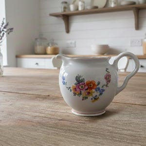 May include: A white ceramic creamer with a floral design in shades of pink, orange, and blue. The creamer has a curved handle and a spout. It sits on a wooden table in a kitchen setting.