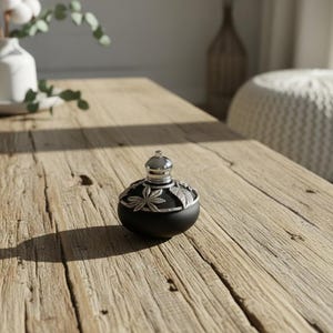 May include: A black decorative jar with a silver lid and leaf accents. The jar sits on a rustic wooden table, with a white vase of cotton stems and a woven pouf in the background. The jar's shadow is cast on the table.