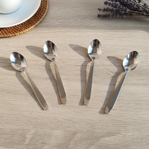 Op de afbeelding: Vier roestvrijstalen lepels liggen op een licht oppervlak met houtnerf. De lepels zijn diagonaal geplaatst en werpen schaduwen. Een witte kop en schotel zijn linksboven te zien, samen met een kleine bundel gedroogde lavendel.