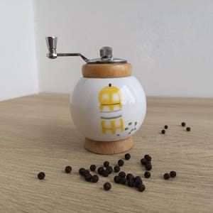 A small, round vintage-style pepper mill with a white porcelain body and a light wooden top. The mill features a subtle yellow geometric graphic on its side and has an exposed metal grinding mechanism at the top. The image is a close-up, showcasing the mill's design and internal structure, resting on a rustic farmhouse wooden table.