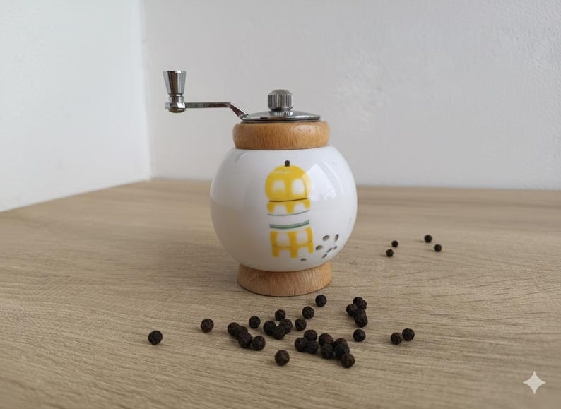 A small, round vintage-style pepper mill with a white porcelain body and a light wooden top. The mill features a subtle yellow geometric graphic on its side and has an exposed metal grinding mechanism at the top. The image is a close-up, showcasing the mill's design and internal structure, resting on a rustic farmhouse wooden table.