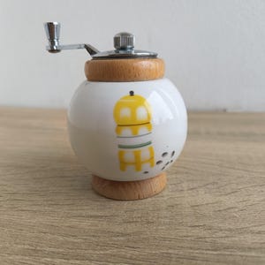 A small, round vintage-style pepper mill with a white porcelain body and a light wooden top. The mill features a subtle yellow geometric graphic on its side and has an exposed metal grinding mechanism at the top. The image is a close-up, showcasing the mill's design and internal structure, resting on a rustic farmhouse wooden table.