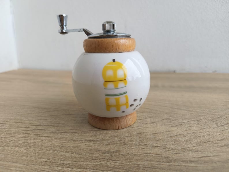 A small, round vintage-style pepper mill with a white porcelain body and a light wooden top. The mill features a subtle yellow geometric graphic on its side and has an exposed metal grinding mechanism at the top. The image is a close-up, showcasing the mill's design and internal structure, resting on a rustic farmhouse wooden table.