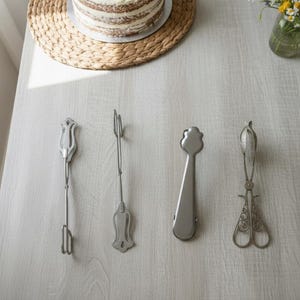 Puede incluir: Cuatro utensilios de servir plateados están dispuestos sobre una superficie de madera gris claro. Un pastel parcialmente visible se encuentra sobre un salvamanteles tejido. También hay un pequeño jarrón de cristal con flores. Los utensilios incluyen un tenedor, una espátula y dos tipos diferentes de pinzas.