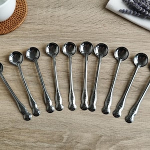 Op de afbeelding: Een verzameling van negen kleine, zilverkleurige lepels met ronde kommen en lange, slanke handvatten. De lepels zijn gerangschikt op een lichtgekleurd houten oppervlak, wat wijst op een keuken- of eetomgeving. Een witte kop en een onderzetter zijn zichtbaar.