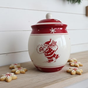 May include: A white and red ceramic cookie jar with a Santa Claus design. The jar has a lid and is decorated with snowflakes. Several snowflake-shaped cookies are arranged on a wooden surface near the jar.