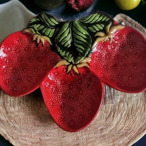 May include: A red ceramic serving dish shaped like three strawberries. The dish has green leaves and a textured surface.