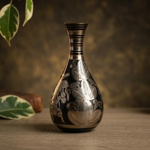 Może przedstawiać: Dekoracyjny czarno-srebrny wazon z grawerunkiem kwiatowym. Wazon ma smukłą szyję i pękaty korpus. Srebrny kwiatowy wzór kontrastuje z czarnym tłem. Wazon jest wykonany z metalu i ma estetykę vintage.