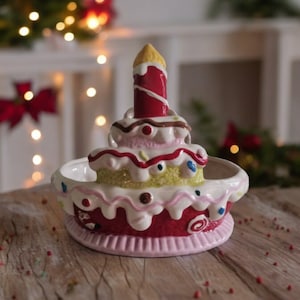 May include: A ceramic cake-shaped dish with a red, white, and yellow glaze. The dish has a single candle on top and is decorated with sprinkles and swirls.