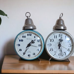 May include: Two vintage alarm clocks with bell tops. The clock on the left has a blue face with black numbers and hands. The clock on the right has a white face with black numbers and hands.