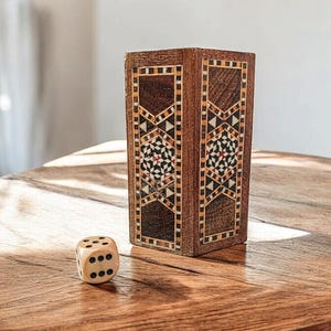 May include: A wooden box with intricate inlaid geometric patterns in shades of brown, red, and white. A single white die with black dots sits on a wooden table in front of the box.