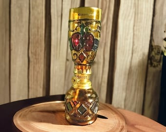 May include: A small, gold-colored oil lamp with a stained glass design featuring red, green, and black accents. The lamp is sitting on a wooden surface.