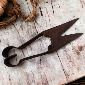 May include: A pair of rusty, antique shears with a unique, heart-shaped handle. The shears are lying on a weathered, white wooden surface.
