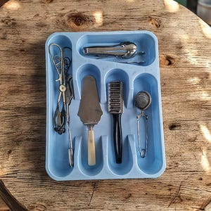 K&ouml;nnte beinhalten: Ein blauer Plastik-Besteckhalter mit sechs F&auml;chern. Der Halter enth&auml;lt einen silbernen Kuchenspachtel, ein silbernes Teesieb, eine silberne B&uuml;rste, eine silberne Zange und einen silbernen Schneebesen.