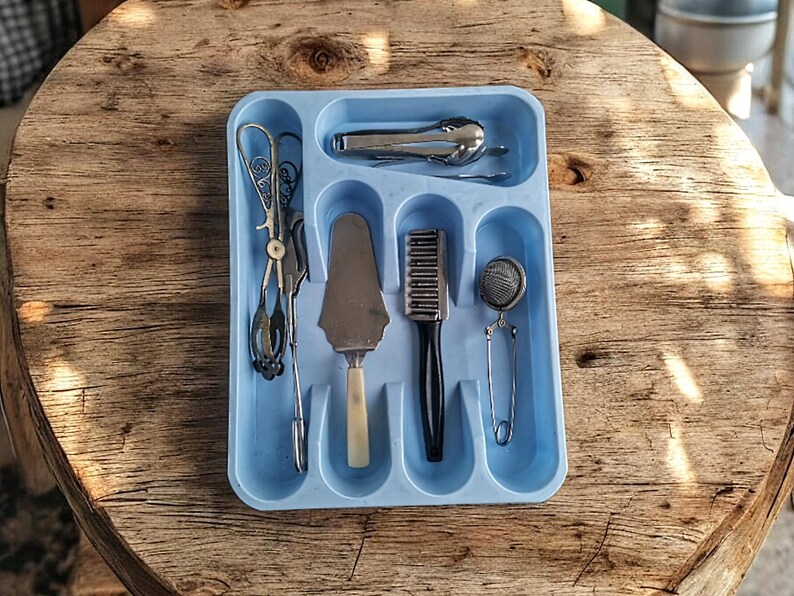 K&ouml;nnte beinhalten: Ein blauer Plastik-Besteckhalter mit sechs F&auml;chern. Der Halter enth&auml;lt einen silbernen Kuchenspachtel, ein silbernes Teesieb, eine silberne B&uuml;rste, eine silberne Zange und einen silbernen Schneebesen.