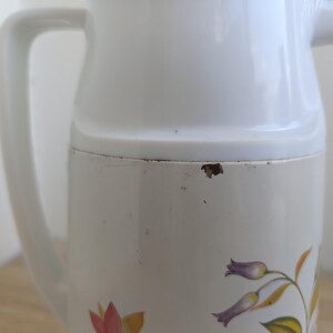 May include: A white vintage pitcher with a handle and a spout. The pitcher has a floral design in shades of purple, yellow, and red. The pitcher is made of a durable material and is suitable for serving beverages.