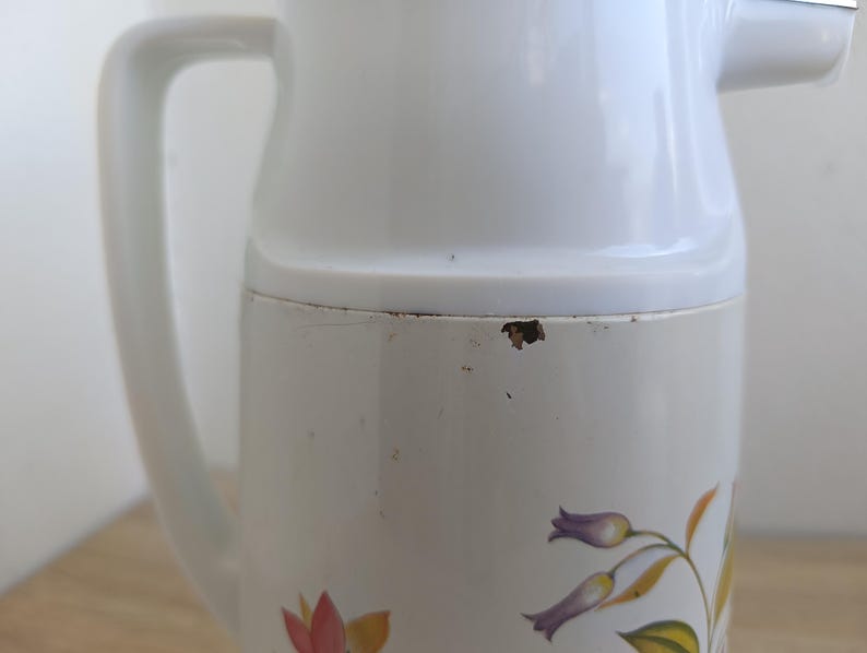 May include: A white vintage pitcher with a handle and a spout. The pitcher has a floral design in shades of purple, yellow, and red. The pitcher is made of a durable material and is suitable for serving beverages.