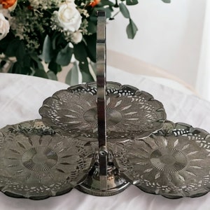 May include: A three-tiered silver serving stand with a handle. Each tier has a scalloped edge and a decorative pattern.