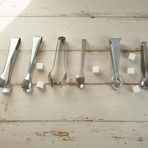 May include: An overhead shot displays six different stainless steel sugar tongs and several sugar cubes on a light-colored wooden surface. The tongs vary in design, with some having claw-like ends and others spoon-shaped.