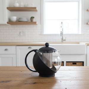 Può includere: Una teiera in vetro trasparente con manico e coperchio neri e un infusore in acciaio inossidabile è appoggiata su un tavolo di legno. Lo sfondo mostra una cucina con mobili bianchi, un lavandino e una finestra.